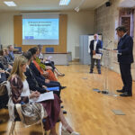 Aula del rectorado de la Universidad de Alcalá. El rector Carmelo García inicia la lectura inclusiva del Quijote delante de un atril y antes el público participante. Al fondo se observa una pantalla donde puede leerse "El Quijote de Cervantes: una lectura inclusiva".
