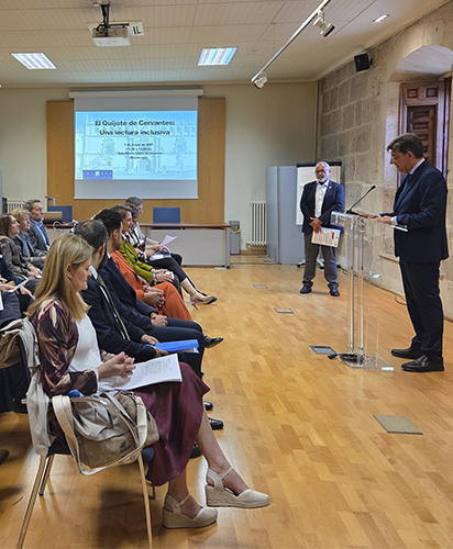 Aula del rectorado de la Universidad de Alcalá. El rector Carmelo García inicia la lectura inclusiva del Quijote delante de un atril y antes el público participante. Al fondo se observa una pantalla donde puede leerse "El Quijote de Cervantes: una lectura inclusiva".