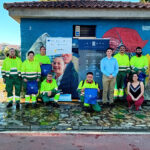 Badajoz web Grupo de personas posando al aire libre junto a una caseta de obra con un mural pintado, en un entorno urbano junto al río Guadiana en Badajoz y zonas verdes. Varias de estas personas llevan uniforme de trabajo de alta visibilidad (chaqueta amarilla y pantalón verde), y algunas sostienen bolsas con el logotipo de Integra CEE. En el centro hay dos roll-ups: uno del programa “Más Empleo Plus” de la Fundación Don Bosco y otro de Integra CEE con el mensaje “Contratamos talento, promovemos inclusión”. En primer término, varias personas están de pie y otras agachadas, formando un grupo.
