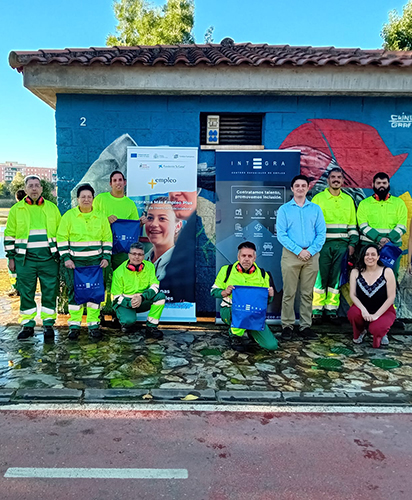 Badajoz web Grupo de personas posando al aire libre junto a una caseta de obra con un mural pintado, en un entorno urbano junto al río Guadiana en Badajoz y zonas verdes. Varias de estas personas llevan uniforme de trabajo de alta visibilidad (chaqueta amarilla y pantalón verde), y algunas sostienen bolsas con el logotipo de Integra CEE. En el centro hay dos roll-ups: uno del programa “Más Empleo Plus” de la Fundación Don Bosco y otro de Integra CEE con el mensaje “Contratamos talento, promovemos inclusión”. En primer término, varias personas están de pie y otras agachadas, formando un grupo.
