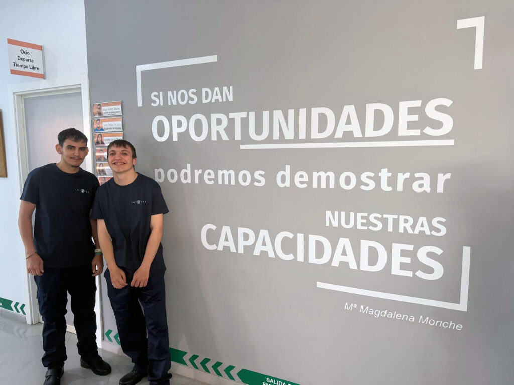 Dos chicos jóvenes con uniforme azul marino y camiseta con el logo de Integra CEE posan en el interior de un edificio junto a una pared con un mensaje en letras grandes: “Si nos dan oportunidades podremos demostrar nuestras capacidades”, firmado por Magdalena Moriche. A un lado se ve una puerta señalizada como “Ocio, Deporte, Tiempo Libre” y paneles con nombres o información.