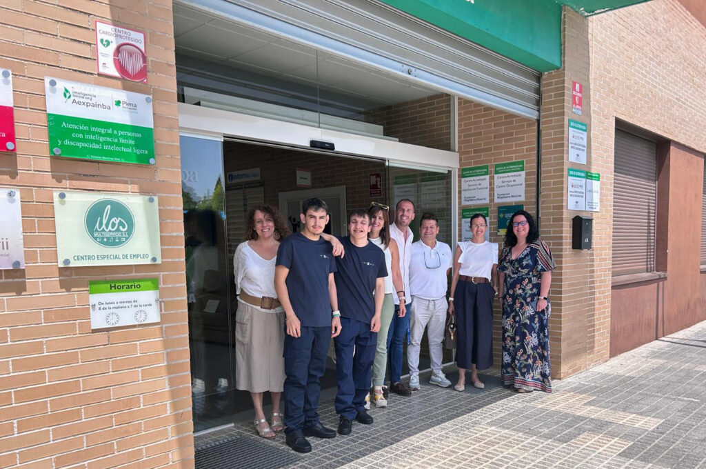 Grupo de personas posando frente a la entrada de un edificio con fachada de ladrillo. Sobre la puerta hay varios carteles, entre ellos uno de “Centro cardioprotegido” y otro de “Aexpainba Plena inclusión”, además de una placa de centro especial de empleo. Varias personas están juntas en la puerta automática, y dos de ellas visten uniforme oscuro. Se aprecia el horario del centro y señalética informativa en la pared.