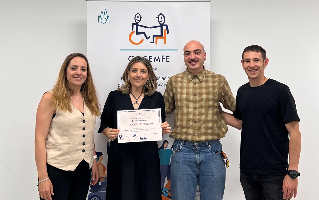 Cuatro personas (dos mujeres y dos hombres) posan frente a un roll-up de COCEMFE. La persona situada en el centro sostiene un certificado en el que se puede leer “Reconocimiento a COCEMFE Burgos”. El resto de participantes se sitúa a ambos lados del diploma en una fotografía de grupo sobre fondo blanco con elementos gráficos relacionados con la discapacidad.