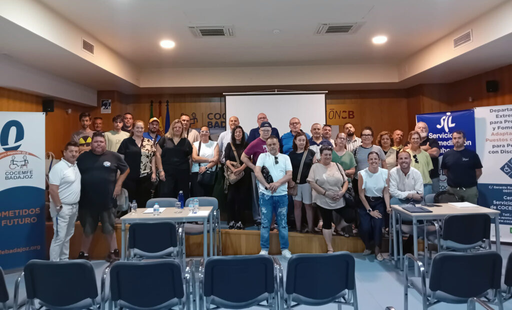 Grupo numeroso de personas posa en el interior de una sala de conferencias, en el marco de la iniciativa "Cambia el rumbo" de Integra CEE. Detrás hay una pantalla de proyección y varias banderas. En los laterales se ven roll-ups de organizaciones, entre ellos Cocemfe Badajoz y servicios relacionados con empleo e inclusión. En primer plano aparecen varias filas de sillas vacías.