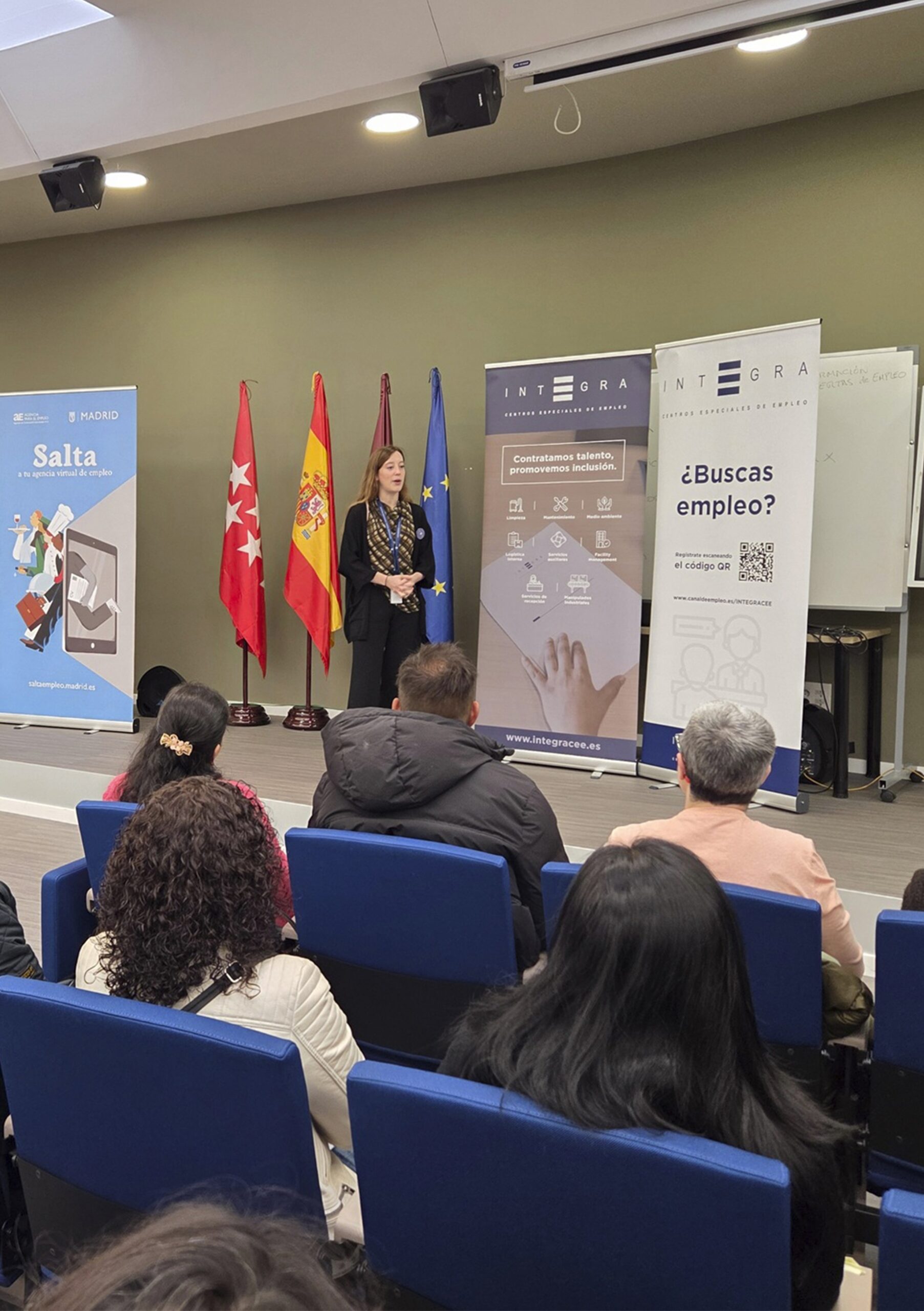 Jornada de "Cambia el rumbo" junto a la Agencia para el Empleo del Ayuntamiento de Madrid. Un persona realizando una presentación en una sala de conferencias ante un grupo de asistentes sentados en butacas azules. Detrás aparecen varias banderas institucionales (Comunidad de Madrid, España y Unión Europea) y dos roll-ups de Integra CEE con mensajes como “Contratamos talento, promovemos inclusión” y “¿Buscas empleo?”. A la izquierda, un cartel de la Agencia para el Empleo del Ayuntamiento de Madrid con el lema “Salta a tu agencia virtual de empleo”.