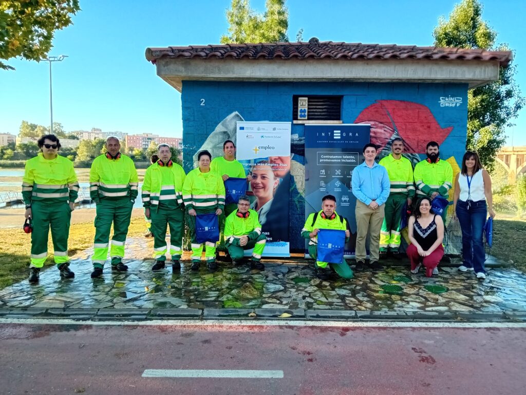 Grupo de personas posando al aire libre junto a una caseta de obra con un mural pintado, en un entorno urbano junto al río Guadiana en Badajoz y zonas verdes. Varias de estas personas llevan uniforme de trabajo de alta visibilidad (chaqueta amarilla y pantalón verde), y algunas sostienen bolsas con el logotipo de Integra CEE. En el centro hay dos roll-ups: uno del programa “Más Empleo Plus” de la Fundación Don Bosco y otro de Integra CEE con el mensaje “Contratamos talento, promovemos inclusión”. En primer término, varias personas están de pie y otras agachadas, formando un grupo. Al fondo se aprecian árboles, edificios y un pequeño pantalán.