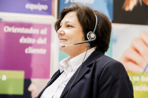 Atención telefónica y servicios auxiliares en Integra CEE Trabajadora de Integra CEE atiende una llamada mediante auriculares con micrófono en un entorno de oficinas, frente a paneles informativos, como parte de sus funciones de recepción y atención telefónica.