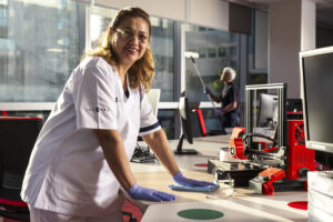 Trabajadora de Integra CEE realiza tareas de limpieza en una oficina con equipamiento tecnológico e impresoras 3D, utilizando guantes de protección y vestuario corporativo; al fondo se aprecia otra persona realizando limpieza profesional de cristales en un ventanal.
