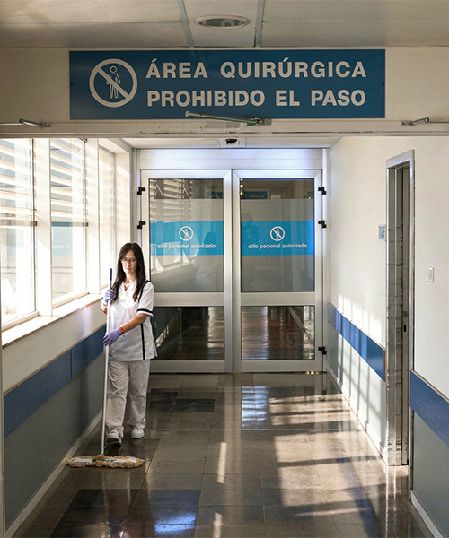 Profesional de Integra CEE realiza tareas de limpieza con mopa en un pasillo interior de un centro hospitalario, junto a puertas de acceso señalizadas con el texto “Área quirúrgica, prohibido el paso” y “solo personal autorizado”. 