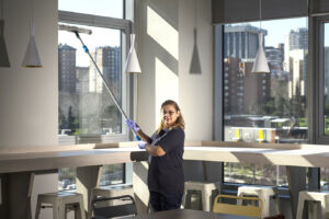 Trabajadora de Integra CEE realiza tareas de limpieza de cristales desde el interior de un espacio luminoso, utilizando una pértiga y elementos de limpieza manual. 