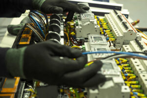 Manipulado de cuadros eléctricos Manos de una persona con guantes de seguridad realizando tareas de manipulado en un cuadro de instalaciones eléctricas, con diferentes piezas y componentes.