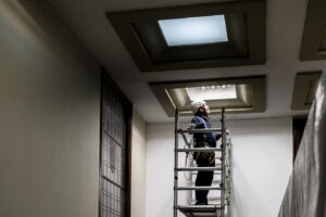 Trabajador de Integra CEE realiza tareas de mantenimiento en altura desde una plataforma de trabajo, utilizando equipos de protección individual, en un espacio interior con iluminación cenital 