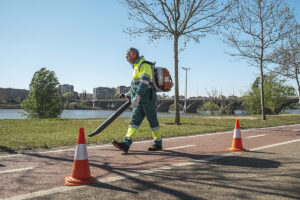 Profesional de Integra CEE realiza tareas de jardinería y limpieza de hojas mediante una sopladora portátil. La zona de trabajo se encuentra señalizada con conos de seguridad, en un parque exterior junto a un espacio fluvial urbano. 