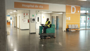 Profesional de Integra CEE subido a una máquina fregadora en el interior de una hospital. Se observa un espacio amplio con señalización, ascensores y mobiliario de espera. Al fondo, el acceso a la zona de hospital de día del centro con un cartel de color naranja. 