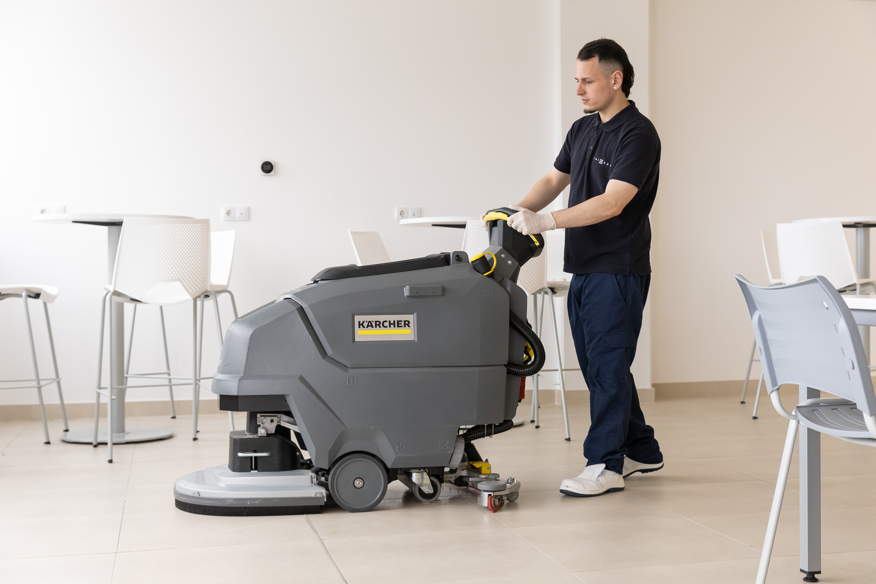Profesional de Integra CEE maneja una fregadora industrial para la limpieza del suelo en un espacio interior diáfano, equipado con mesas y sillas altas, utilizando vestuario corporativo y guantes y calzado de protección.