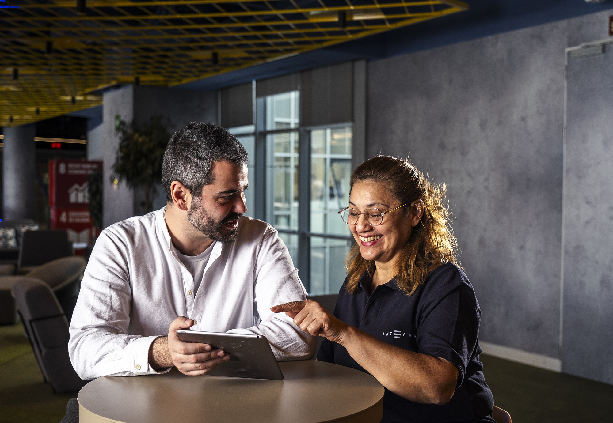 Técnico de apoyo y trabajadora de Integra CEE en una sesión de acompañamiento sociolaboral. Las dos personas se encuentran sentadas alrededor de una mesa alta redonda en el interior de un espacio moderno de oficinas.
