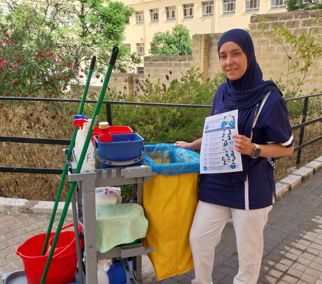 Persona trabajadora del servicio de limpieza posa junto a un carro de limpieza en una zona exterior ajardinada del IES Siete Colinas de Ceuta. En una mano sostiene un folleto informativo de la campaña "Comunicar es conectar, conectar es incluir", mientras detrás se observan vegetación, un edificio y un camino pavimentado.