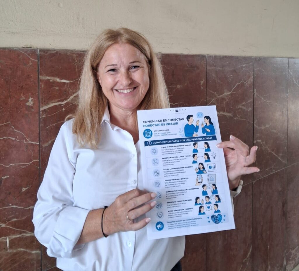 Persona trabajadora de Integra CEE muestra un cartel de la campaña "Comunicar es conectar, conectar es incluir" ante una pared revestida de mármol del Acueartelamiento de la Marina de Ceuta. El cartel reúne pautas para favorecer una comunicación accesible con personas sordas y fomentar la inclusión en el entorno laboral.