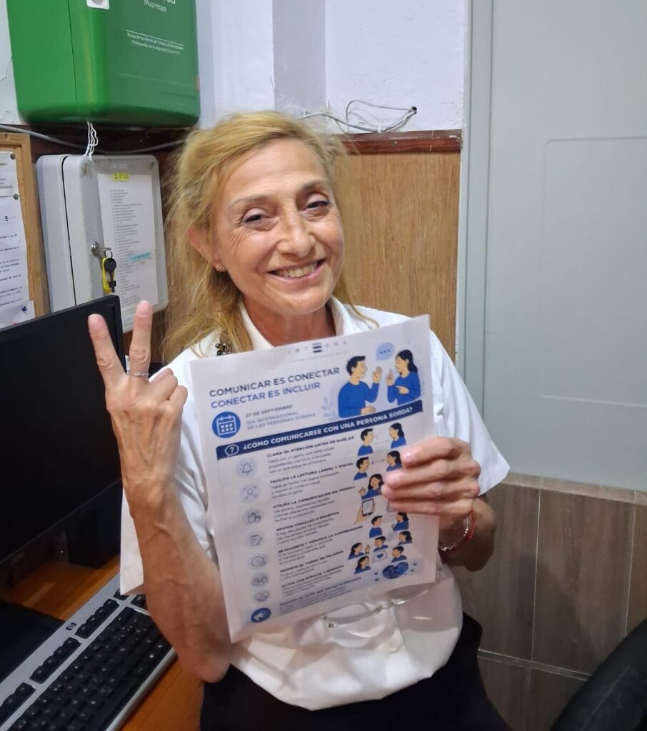 Persona trabajadora de Integra CEE muestra un cartel de la campaña "Comunicar es conectar, conectar es incluir" en un espacio interior del Centro Deportivo y Cultural Militar de Ceuta. El cartel recoge recomendaciones para facilitar la comunicación con personas sordas y fomentar entornos más inclusivos.