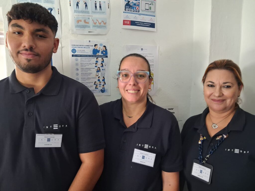 Tres personas trabajadoras de Integra CEE posan en un espacio de las instalaciones de los Jardines de la Hípica, en Ceuta, y delante de varios carteles informativos colocados en la pared. Entre ellos destaca una lámina informativa de la campaña "Comunicar es conectar, conectar es incluir", con recomendaciones para facilitar la comunicación con personas sordas y promover entornos más inclusivos.