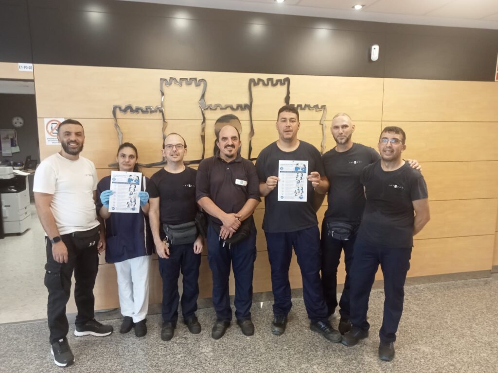 Grupo de personas trabajadoras de Integra CEE posan en el interior del centro educativo IES Almina, emplazado en Ceuta, junto a carteles de la campaña "Comunicar es conectar, conectar es incluir". Dos de las personas sostienen materiales informativos con recomendaciones para facilitar la comunicación con personas sordas y promover entornos más inclusivos.