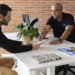 Interior de una oficina con una pared de ladrillo visto. Dos personas mantienen una conversación en torno a una mesa blanca mientras revisan documentación. Una de ellas entrega una hoja de papel a la otra. Sobre la mesa hay una planta de interior, un monitor de ordenador y varios folletos y documentos, en el marco de colaboración entre Integra CEE y Agifes.
